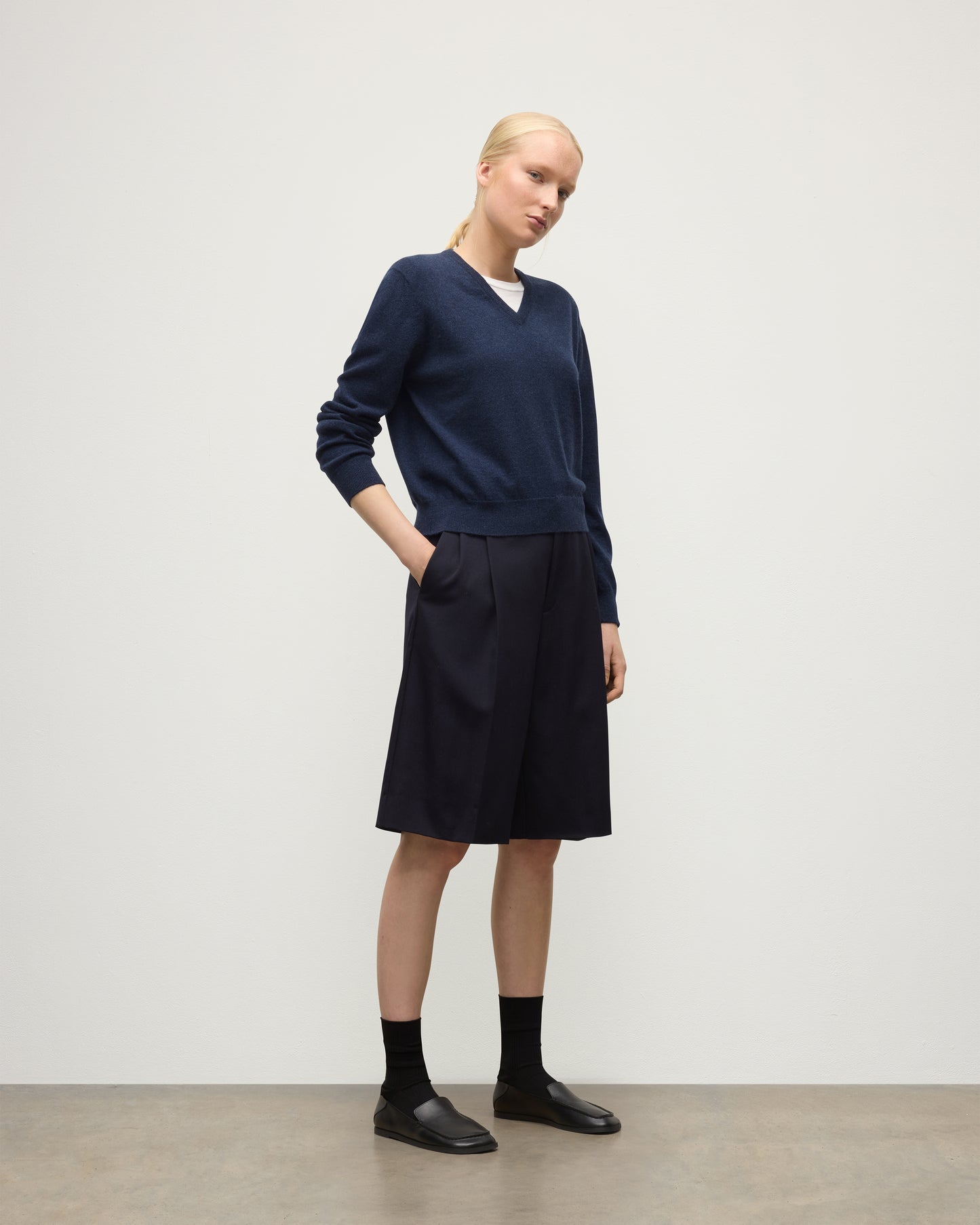 MODEL WEARING JOHNSTONS OF ELGIN WOMENS LUNAN CLASSIC VEE SWEATER NAVY 100% CASHMERE WITH LONG BLACK SHORTS KAP05807HD7350
