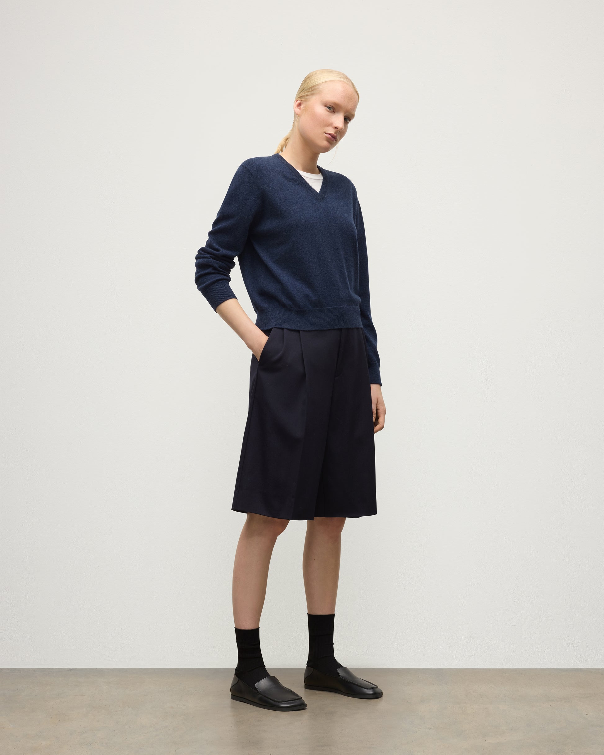 MODEL WEARING JOHNSTONS OF ELGIN WOMENS LUNAN CLASSIC VEE SWEATER NAVY 100% CASHMERE WITH LONG BLACK SHORTS KAP05807HD7350
