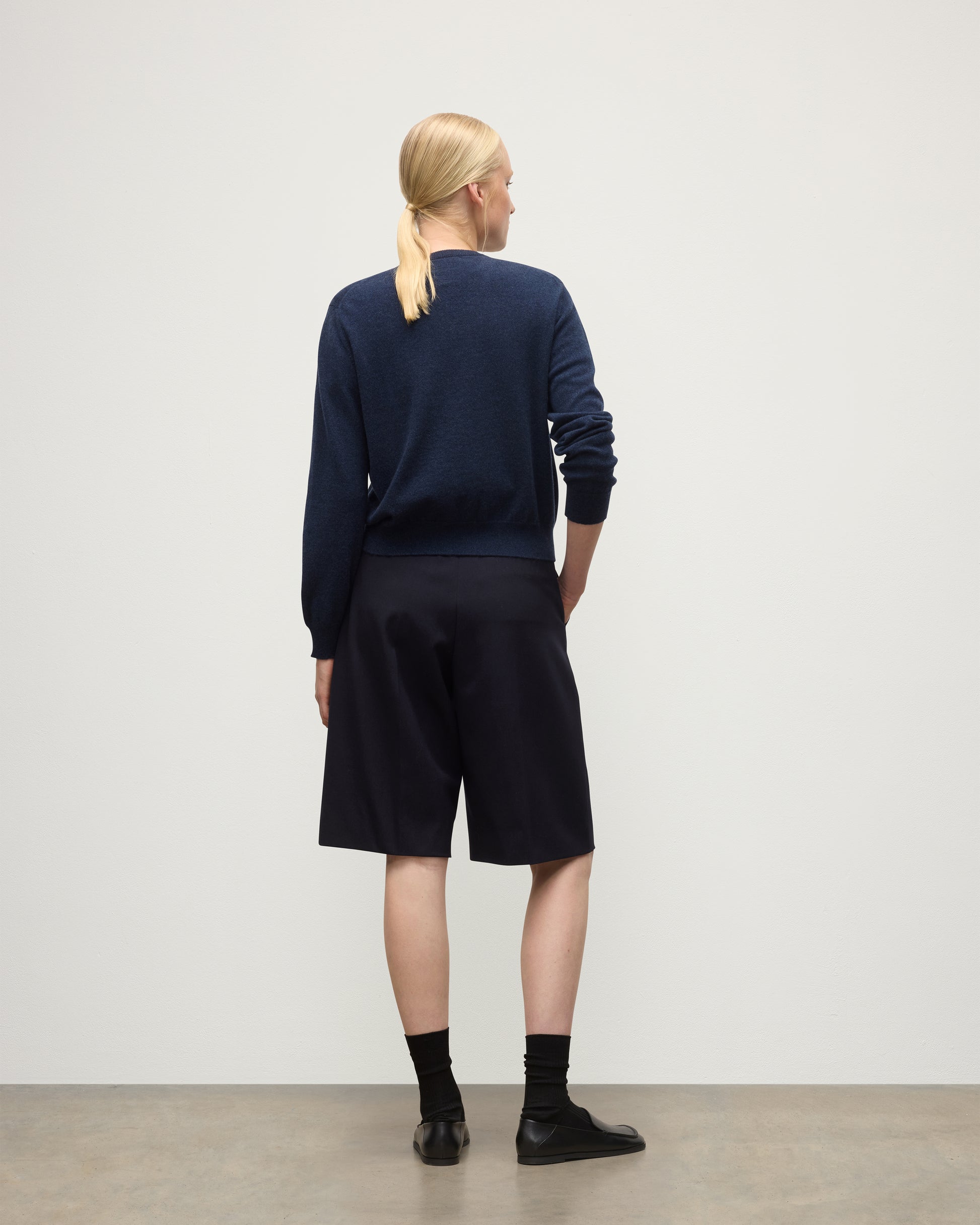 MODEL WEARING JOHNSTONS OF ELGIN WOMENS LUNAN CLASSIC VEE SWEATER NAVY 100% CASHMERE WITH LONG BLACK SHORTS KAP05807HD7350
