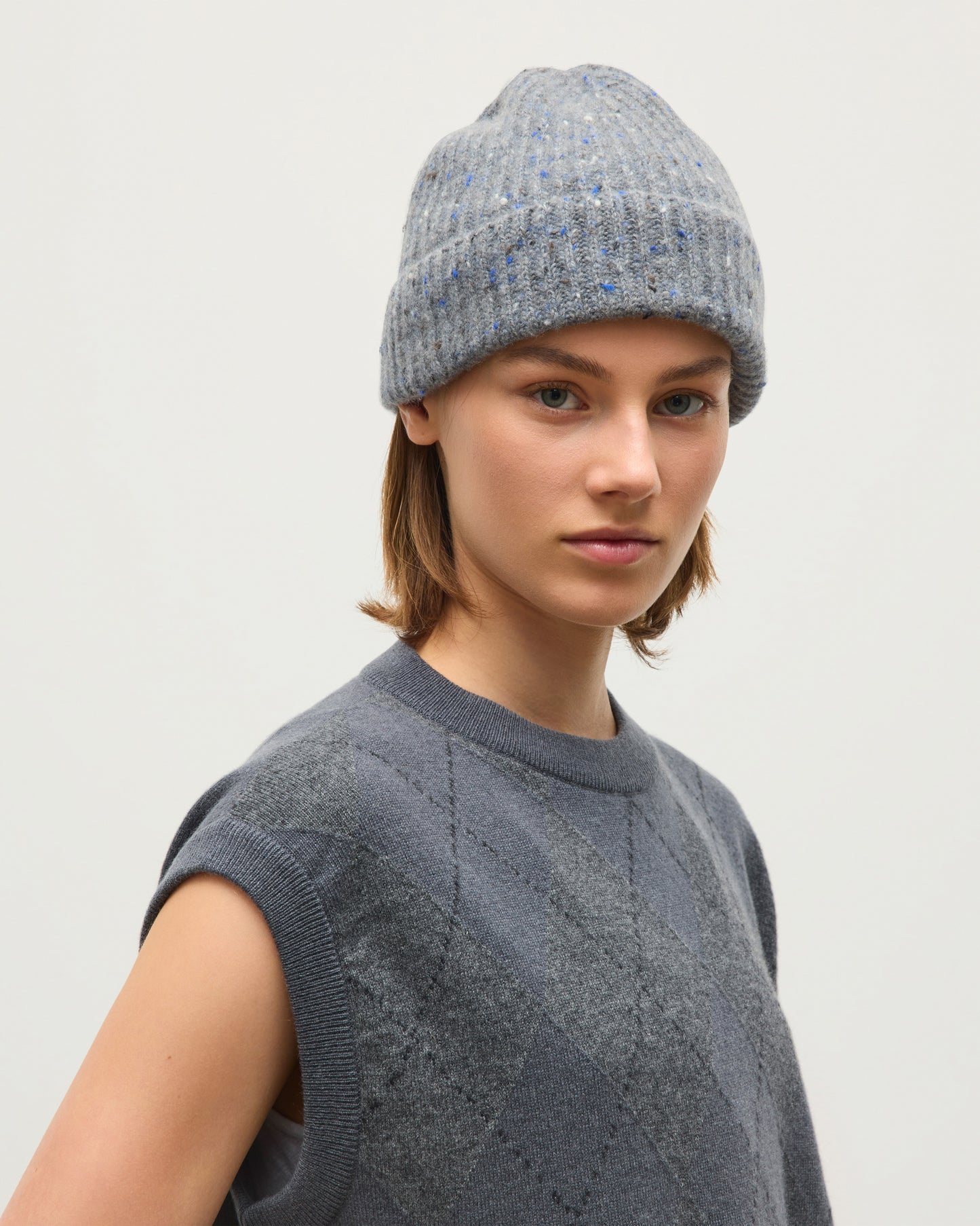 A model wearing a light gray ribbed knit beanie with subtle blue and white flecks, paired with a matching gray sleeveless knit sweater vest, against a plain light gray background. JOHNSTONS OF ELGIN CLAN SLEEVELESS CREW CASHMERE ARGYLE INTARSIA ROUND NECK TANK - 2 PLY GREY 100% CASHMERE KAP06027JU1653