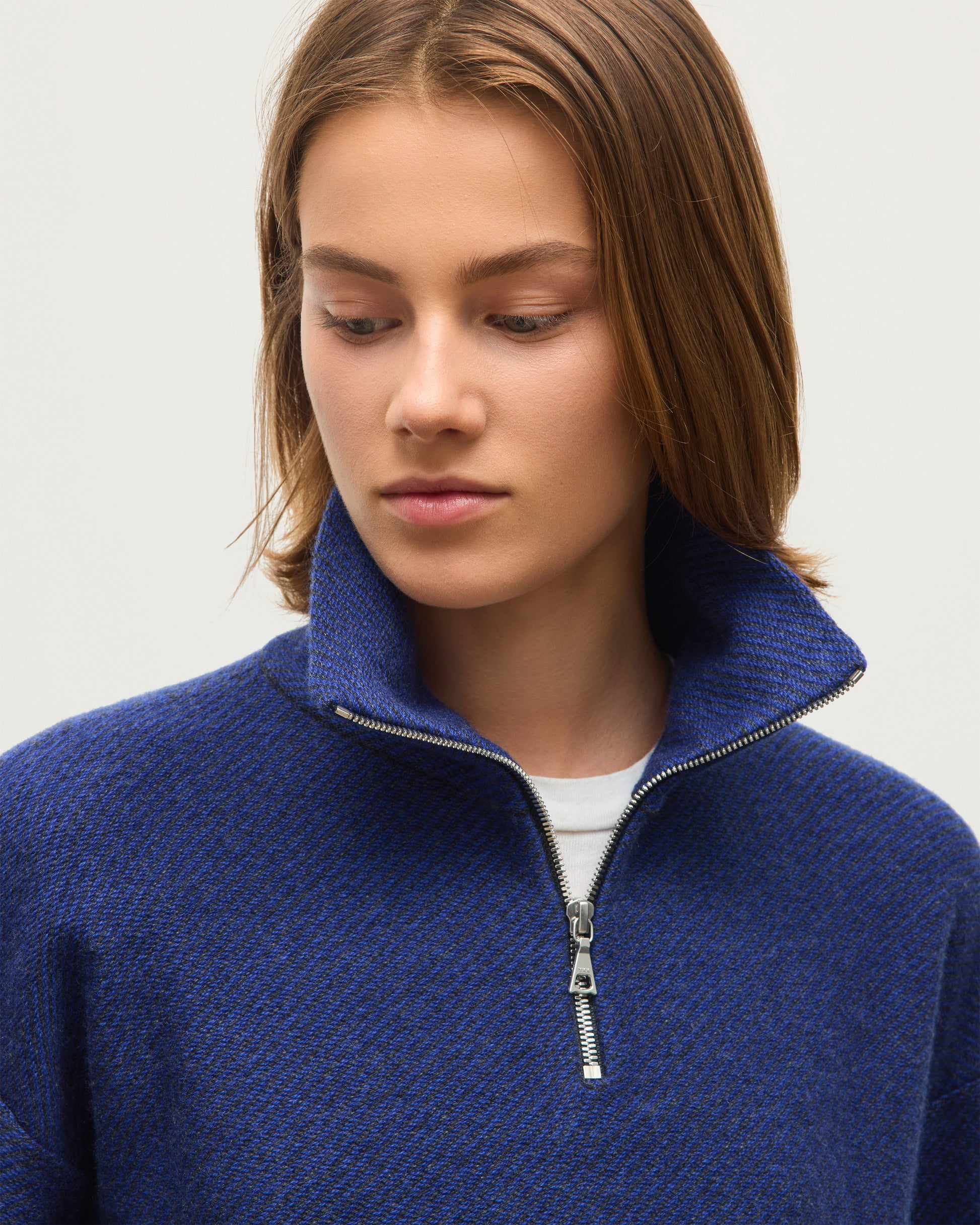 A close-up of a woman wearing a textured blue half-zip sweater with a high collar, layered over a white undershirt, against a plain light gray background. JOHNSTONS OF ELGIN REEL ZIP SWEATER CASHMERE TWILL STITCH ZIP NECK SWEATER - 2 PLY GREY & BLUE 100% CASHMERE KAP06030Q26336