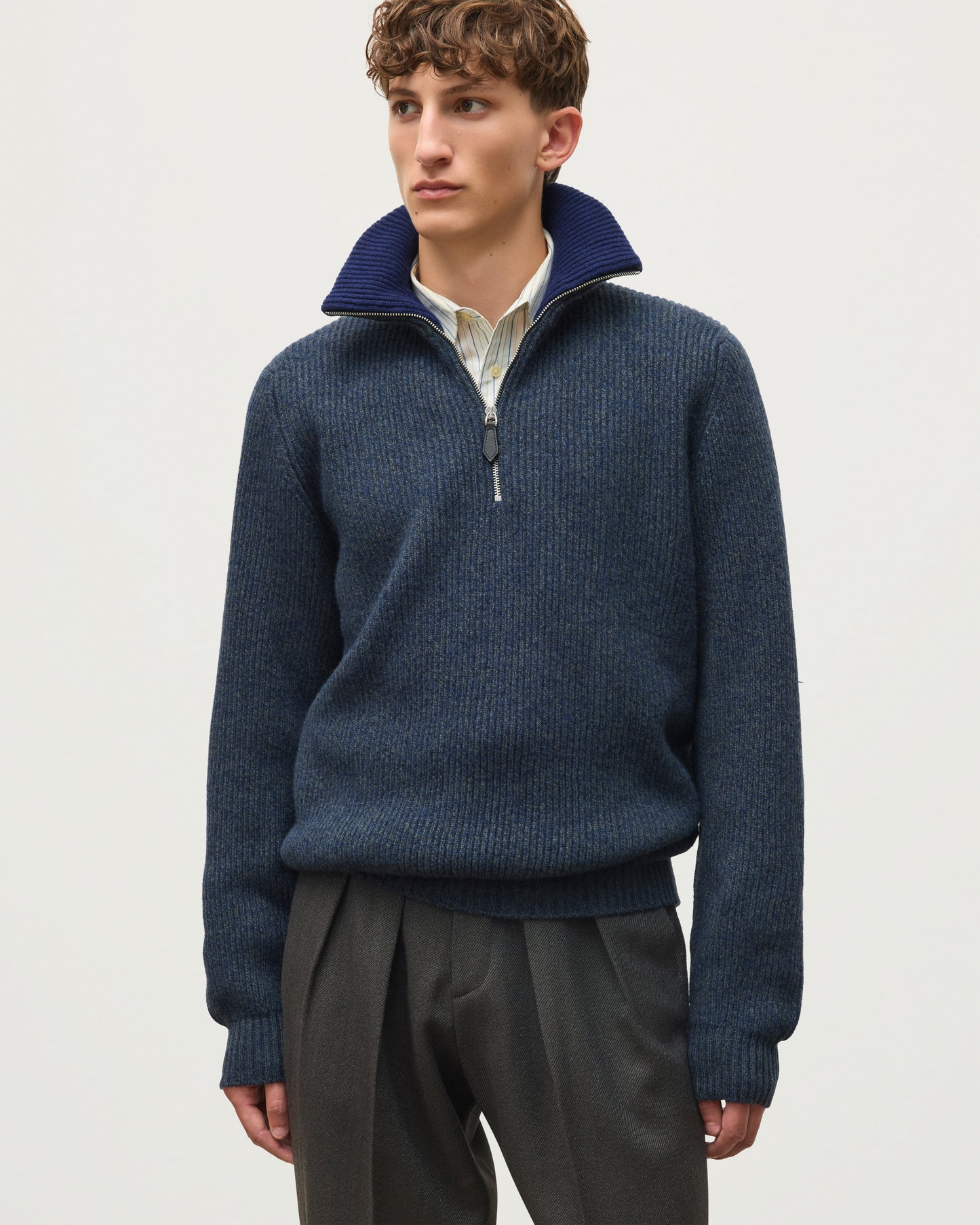 A man wearing a navy blue ribbed knit quarter-zip sweater with a stand-up collar, layered over a light-colored collared shirt and paired with dark gray trousers, photographed against a plain light gray studio background. JOHNSTONS OF ELGIN CHALMERS HALF ZIP SWEATER LAMBSWOOL RIBBED HALF ZIP SWEATER BLUE 100% LAMBSWOOL KDA00819Q26329