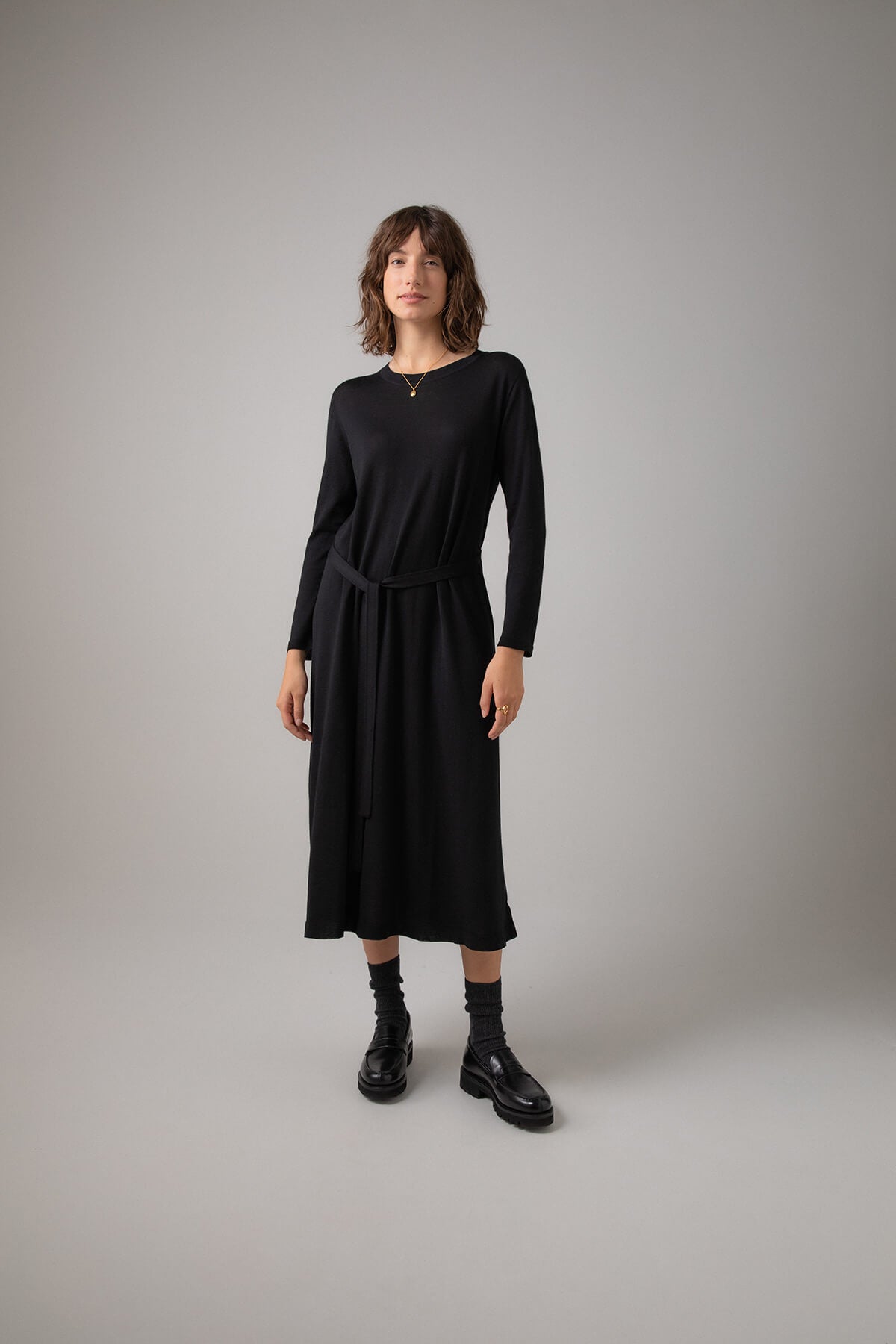 Johnstons of Elgin Women's Superfine Merino Belted T-Shirt Dress in Black worn with Black Socks & Shoes on a grey background KDI00685SA7131