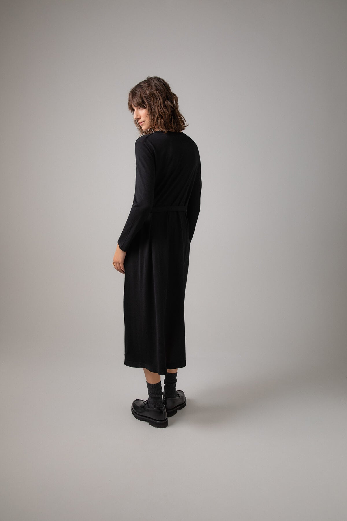 Back of Johnstons of Elgin Women's Superfine Merino Belted T-Shirt Dress in Black worn with Black Socks & Shoes on a grey background KDI00685SA7131