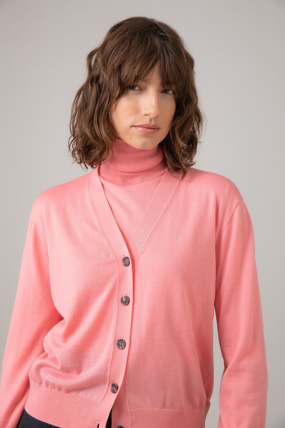 Johnstons of Elgin Women's Superfine Merino Cardigan in Orkney Pink worn with a matching Roll Neck on a grey background KDI00686SE4947