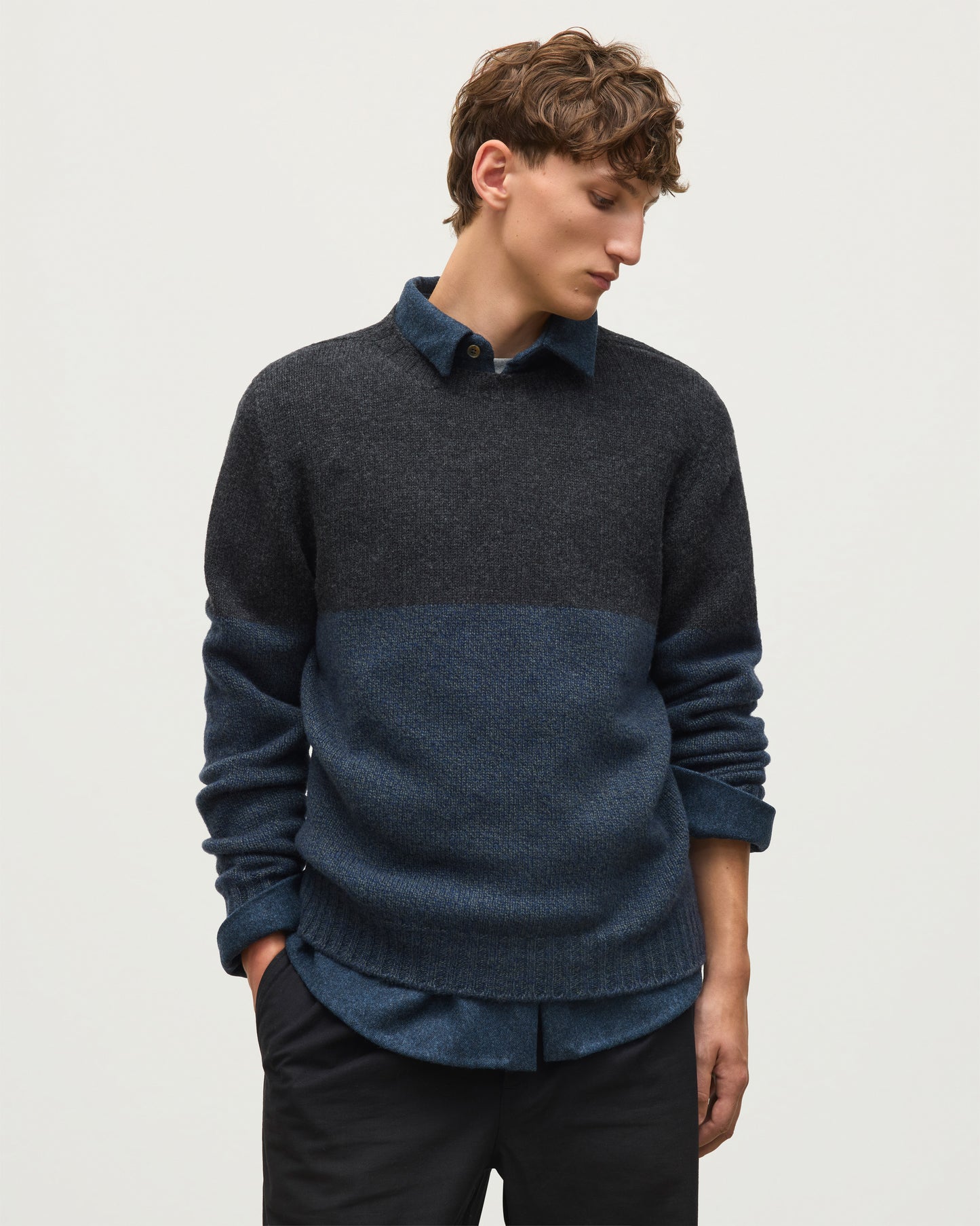 JOHNSTONS OF ELGIN OGILVIE CREW SWEATER LAMBSWOOL COLOURBLOCK CREW NECK SWEATER DARK GREY 100% LAMBSWOOL A man wearing a dark charcoal and navy blue color-block knit sweater over a matching navy collared shirt, paired with black trousers, against a plain light gray background. KDI00816Q26348 