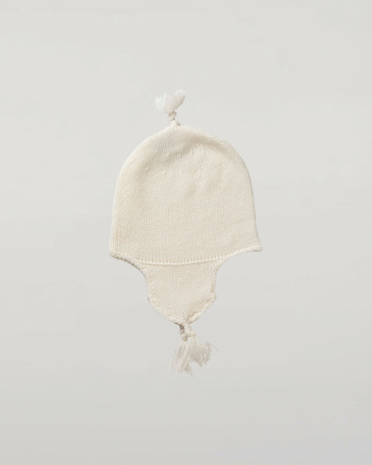 An off-white knit baby hat with ear flaps and tassels, photographed against a neutral light gray Johnstons of Elgin Baby Handknits Ecru Cashmere Baby Hat with Tassel JA000139SA0132ONE