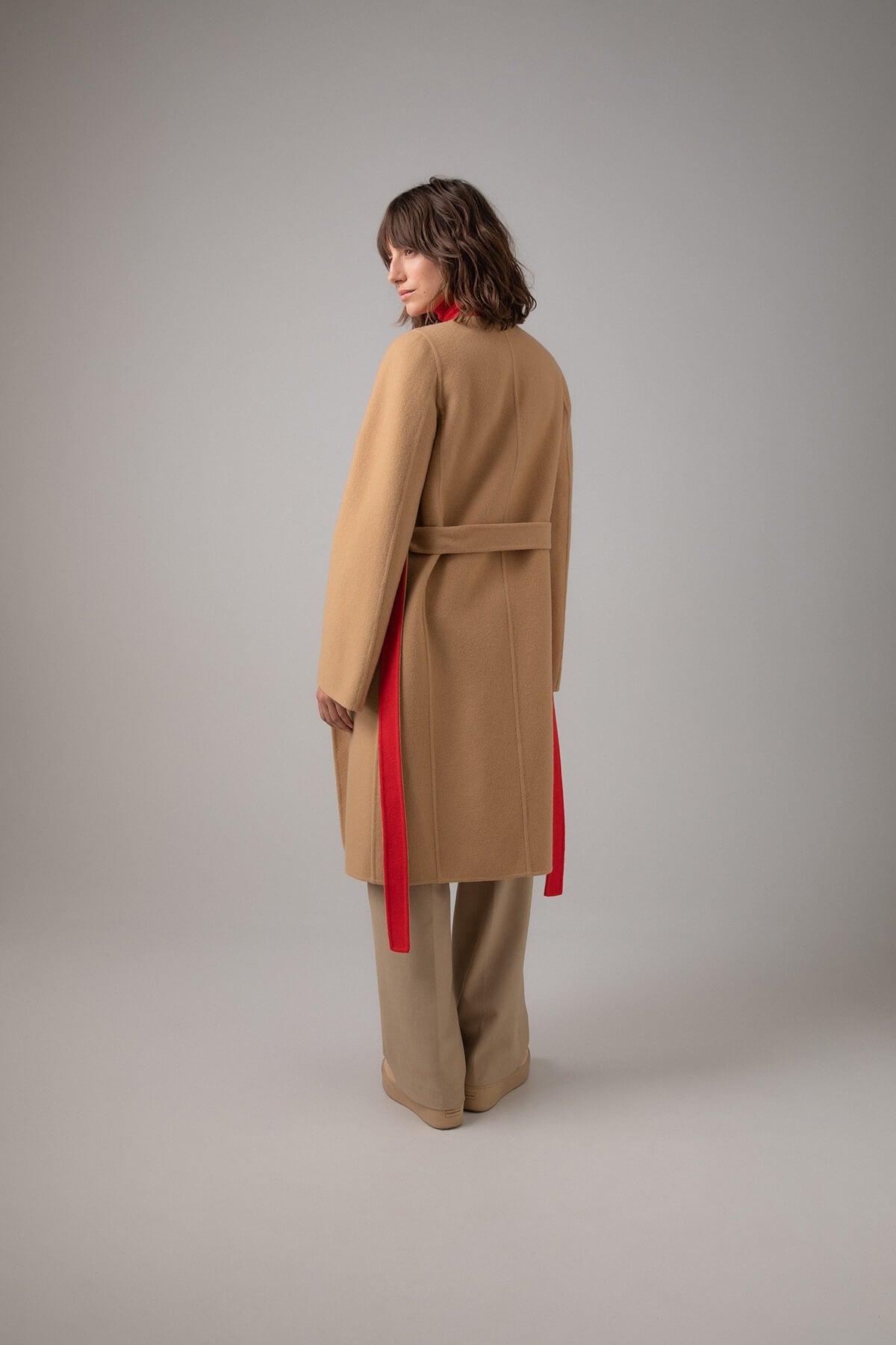 Back of Johnstons of Elgin Women's Collarless Cape Wool Blend Coat in Camel & Red worn with a matching Roll Neck Sweater and Camel Trousers on a grey background TB000633RU7380