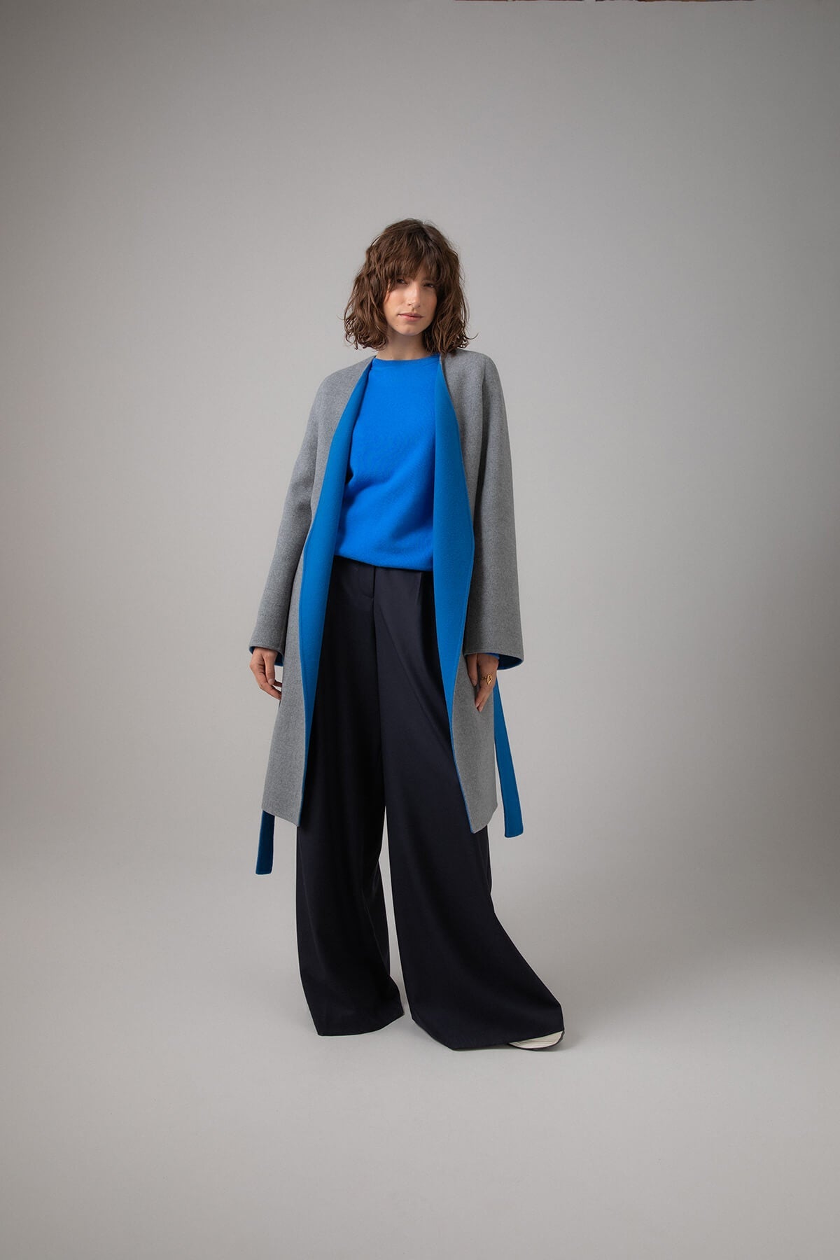 Model wearing Johnstons of Elgin Women's Collarless Cape Wool Blend Coat in Mid Grey & Blue worn with an Orkney Blue Sweater and Navy Trousers on a grey background TB000633RU7381