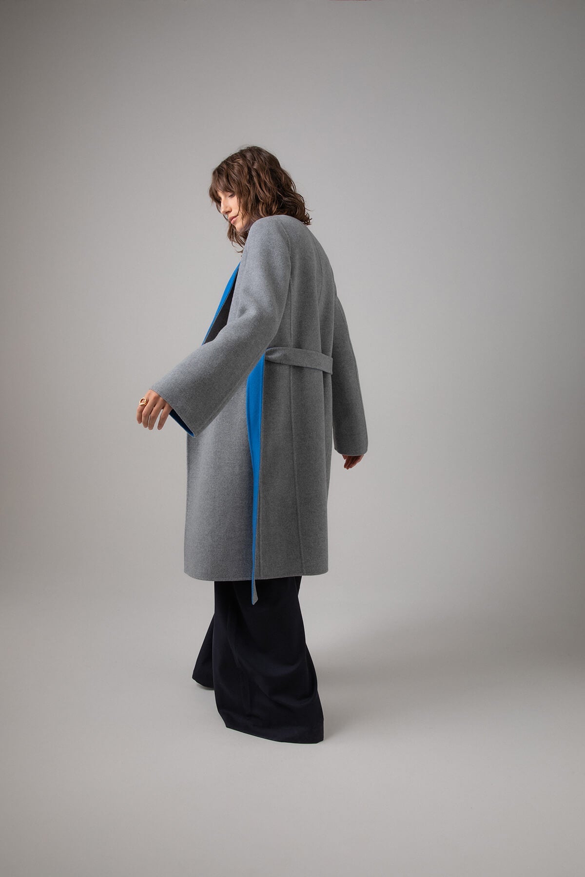 Back of Johnstons of Elgin Women's Collarless Cape Wool Blend Coat in Mid Grey & Blue worn with an Orkney Blue Sweater and Navy Trousers on a grey background TB000633RU7381