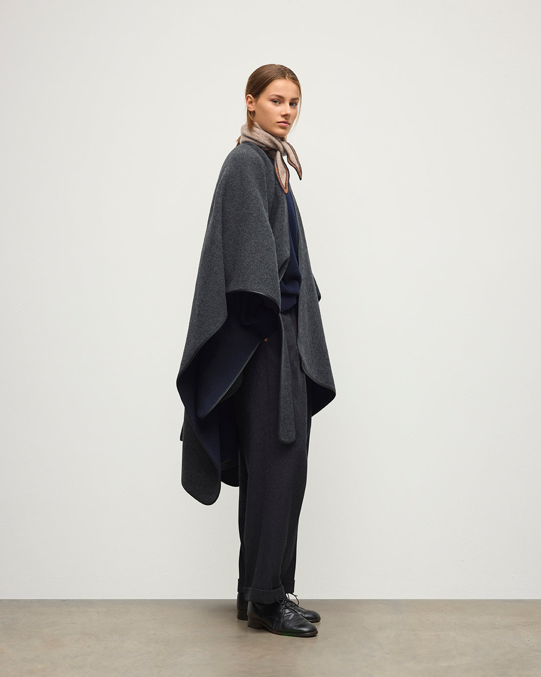A model stands in profile wearing a dark gray draped cape-style outer layer over a navy top and dark trousers, with a beige scarf and black shoes, against a plain light gray studio background. JOHNSTONS OF ELGIN WOMENS BELTED CAPE WITH LEATHER BINDING CHARCOAL & NAVY 90% WOOL, 10% CASHMERE TB000696RU7825ONE
