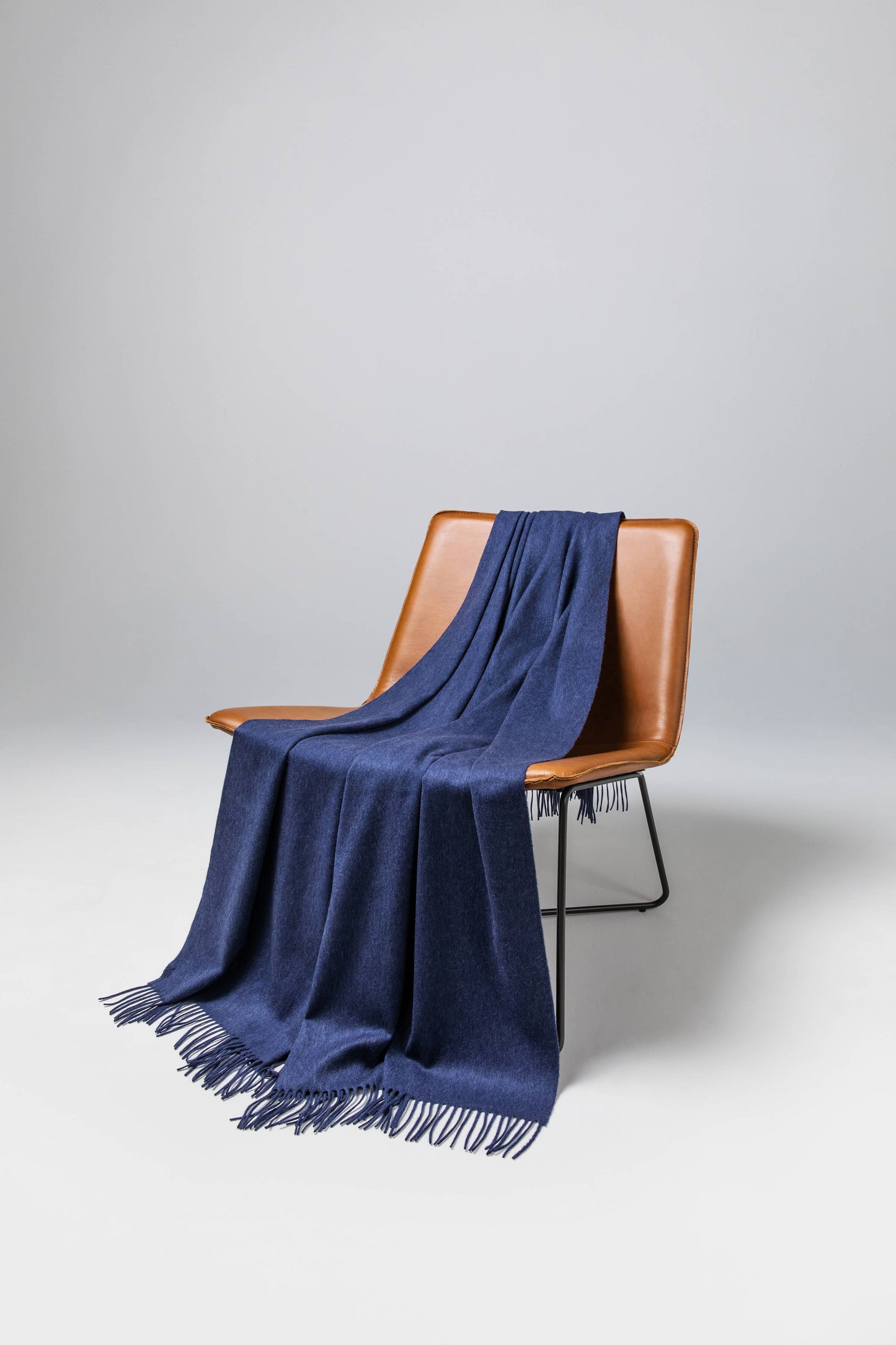 Johnstons of Elgin’s Denim Blue Cashmere Throw on brown chair on a grey background WA000055HD0724