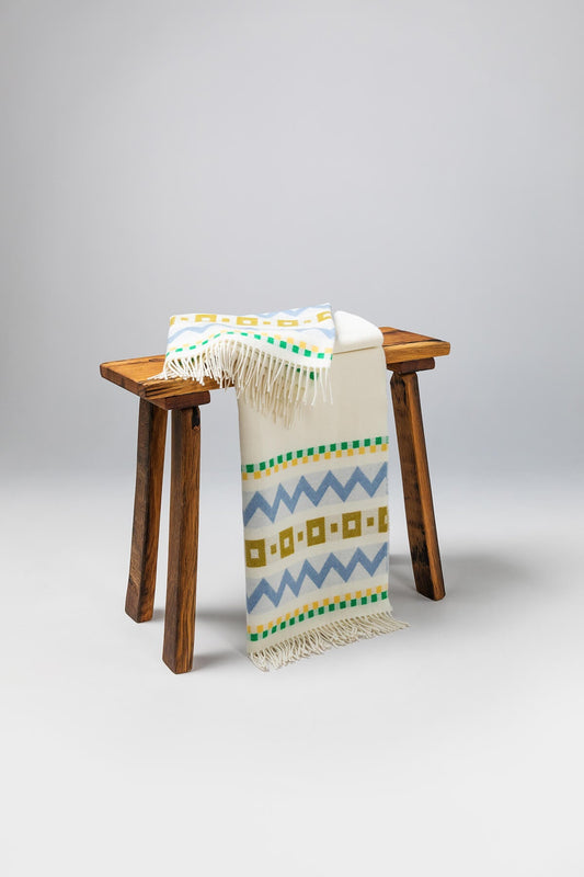 Draped over a small wooden stool a Johnstons of Elgin Children's Zig Zag Blanket in shades of blue, green, and cream on a soft grey background WB002334RU7276ONE