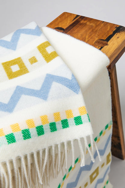  A small wooden stool with a Johnstons of Elgin Children's Zig Zag Blanket in shades of blue, green, and cream on a soft grey background WB002334RU7276ONE