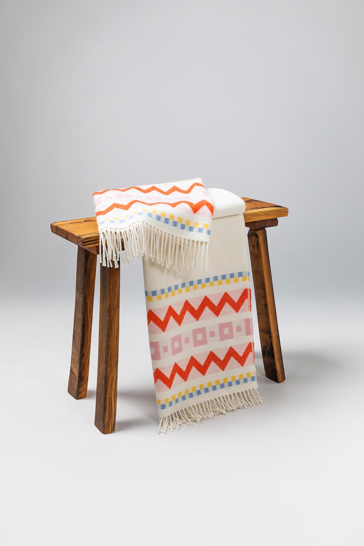 Draped over a small wooden stool a Johnstons of Elgin Children's Zig Zag Blanket in shades of pink, orange, and cream on a soft grey background. WB002334RU7277ONE