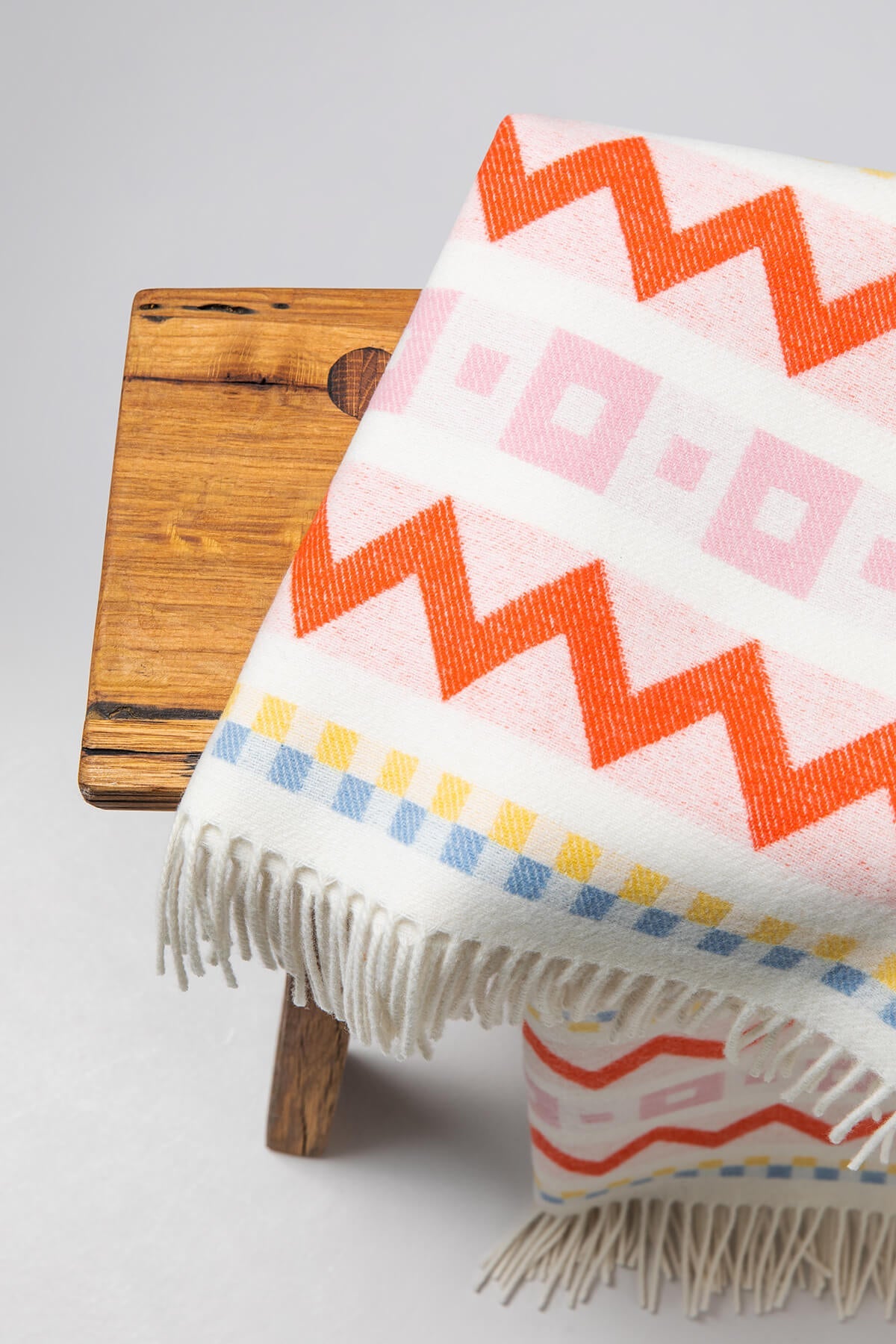  A small wooden stool with a Johnstons of Elgin Children's Zig Zag Blanket in shades of pink, orange, and cream on a soft grey background. WB002334RU7277ONE