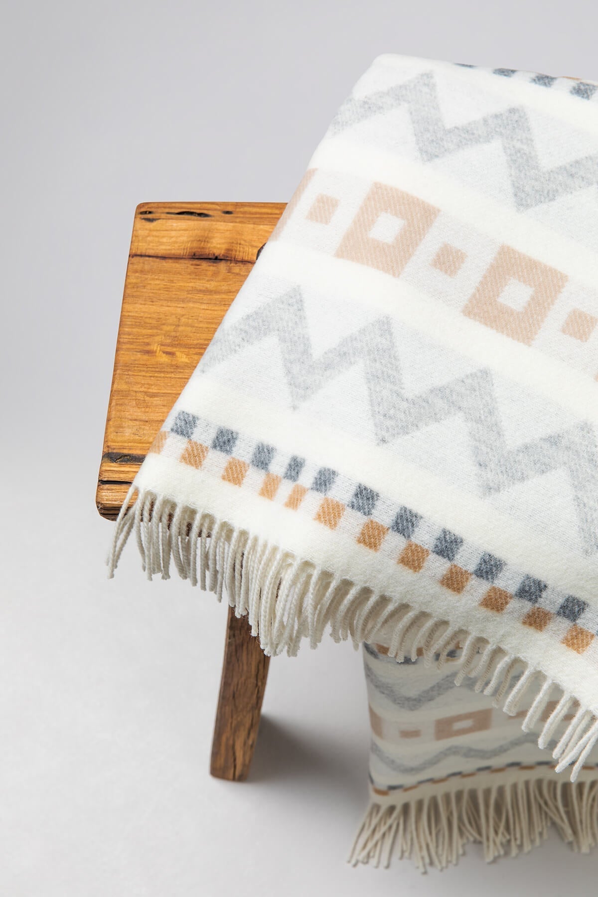 A small wooden stool with a Johnstons of Elgin Children's Zig Zag Blanket in shades of grey, camel, and cream on a soft grey background.WB002334RU7278ONE