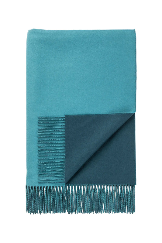 Johnstons of Elgin’s Teal Blue & Ocean Reversible Merino Blend Throw on brown chair on a grey background WB000751RU7260ONE