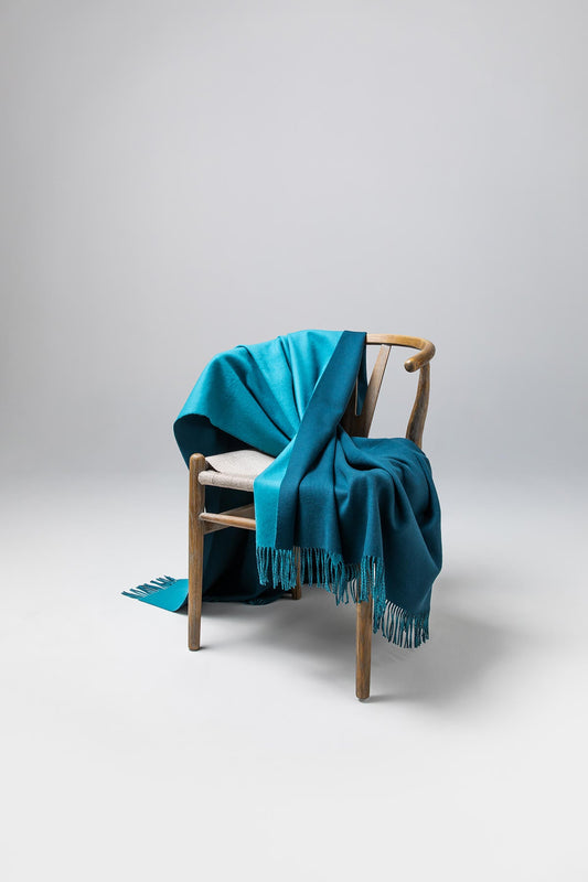 Johnstons of Elgin’s Teal Blue & Ocean Reversible Merino Blend Throw on brown chair on a grey background WB000751RU7260ONE