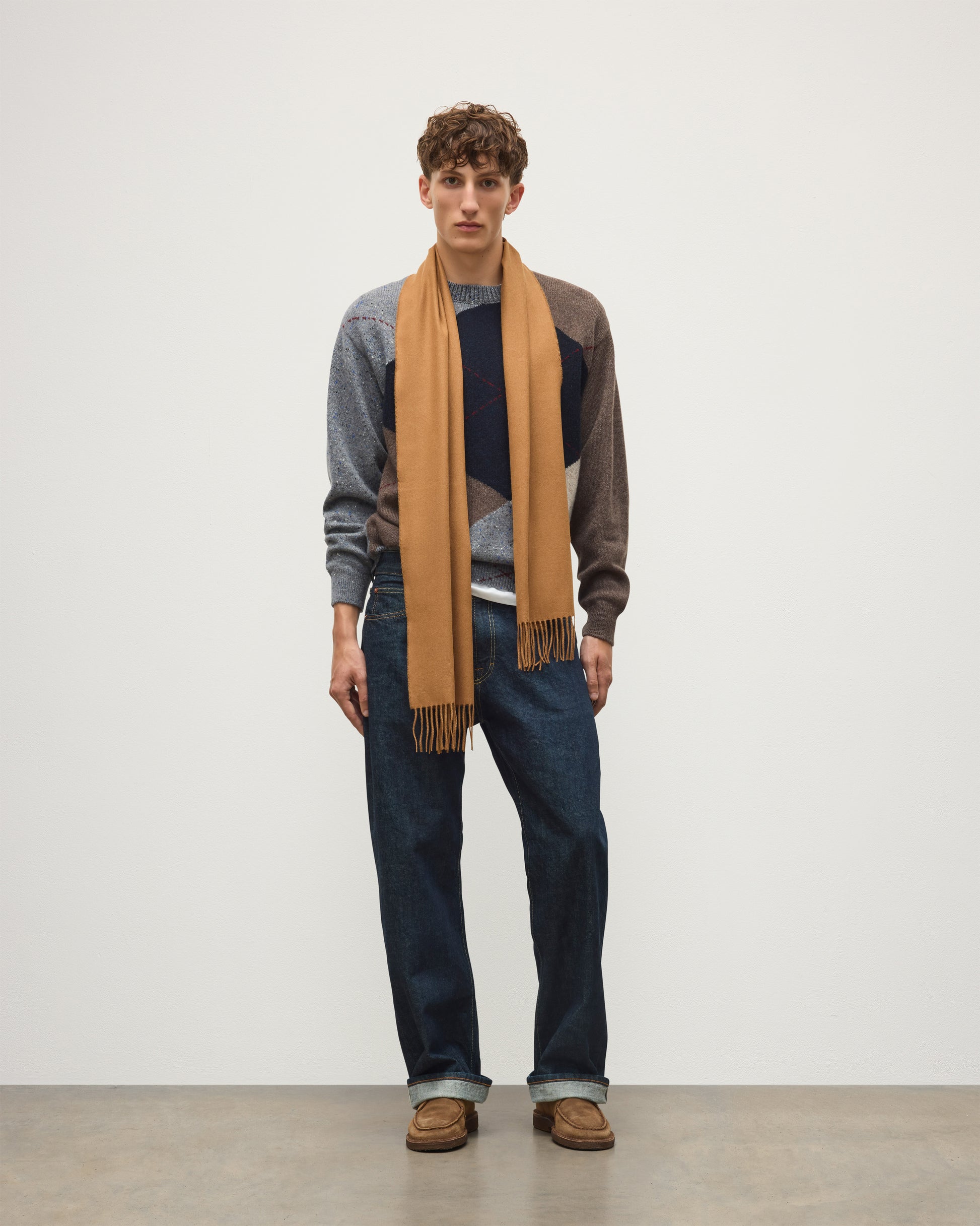A man stands against a plain light gray studio wall wearing a tan scarf draped over a gray and brown color-block sweater, dark blue jeans, and brown shoes. Johnstons of Elgin Vicuna  Pure Vicuña Scarf WR000025SB3022ONE