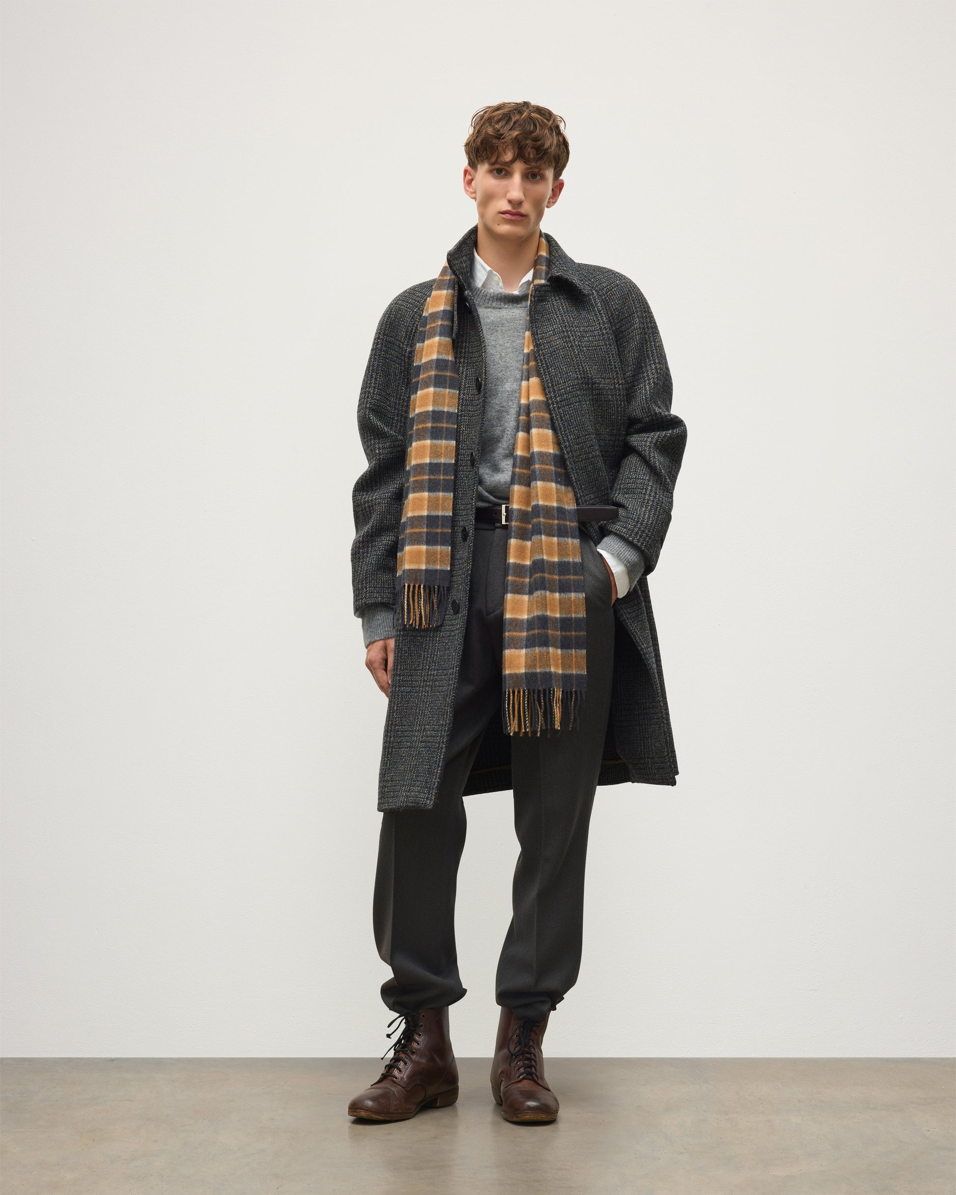 A male model stands against a plain light gray studio background wearing a dark charcoal overcoat, a gray sweater, a brown-and-cream plaid scarf, dark trousers, and brown leather boots. Johnstons of Elgin Vicuna  Windowpane Check Cashmere & Vicuña Scarf WV000013RU6366N/A