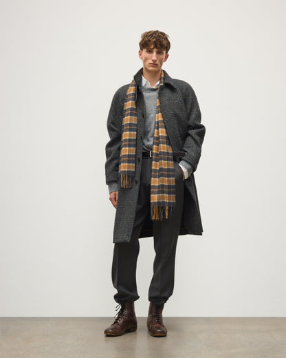 A male model stands against a plain light gray studio background wearing a dark charcoal overcoat, a gray sweater, a brown-and-cream plaid scarf, dark trousers, and brown leather boots. Johnstons of Elgin Vicuna  Windowpane Check Cashmere & Vicuña Scarf WV000013RU6366N/A