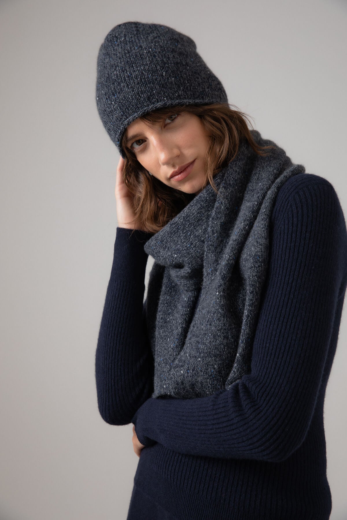 Johnstons of Elgin’s Mid Grey Cashmere Donegal Marl Beanie on model wearing cashmere grey scarf with ribbed navy cashmere jumper on a grey background  HAY03303004529