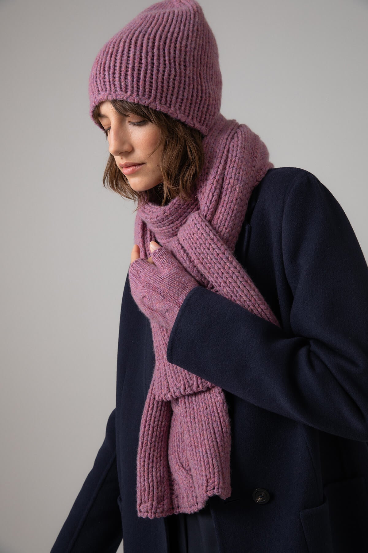 Johnstons of Elgin’s Heather pink Chunky Jersey Cashmere Hat on model wearing matching cashmere scarf and wrist warmers with navy coat HAB03196HE4307