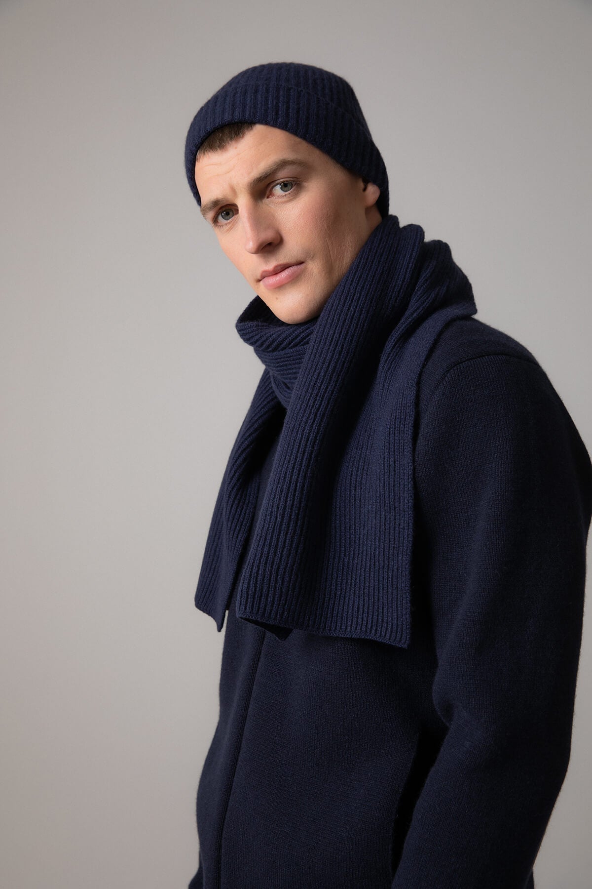 Johnstons of Elgin’s Ribbed Cashmere Beanie and Scarf Giftset in Navy on a model wearing a navy cashmere zip cardigan on a grey background AW23GIFTSET6B